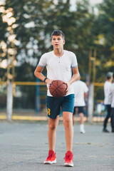 Cute boy in white t shirt plays basketball on a city playground. Active teen enjoying outdoor game with orange ball. Hobby, active lifestyle, sport for kids.	