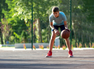 Cute boy in green t shirt plays basketball on a city playground. Active teen enjoying outdoor game with orange ball. Hobby, active lifestyle, sport for kids.	