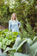 young pretty woman with blue shirt and gloves with flower design posing by raised bed full of fresh vegetables and lettuce