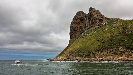 A picturesque rock juts out into the Atlantic Ocean. Steep rocky slopes with sparse vegetation. The boat floats on the water. There are dense clouds in the sky. Cape Town. South Africa