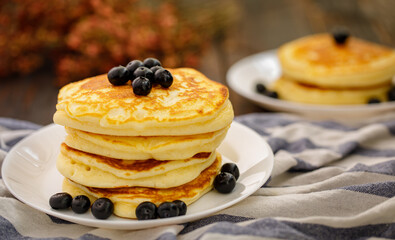 Stack of delicious pancakes sweet food with blueberries and blur dry leaves, plaid fabric in white plate on wooden background.