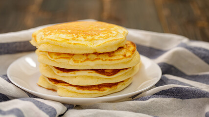 Stack of delicious pancakes sweet food on plaid fabric in white plate on wooden background.