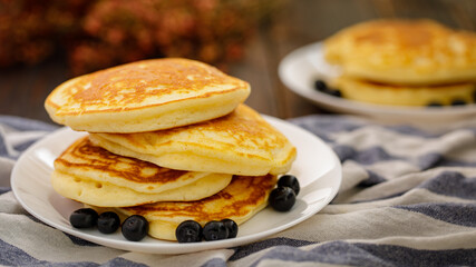 Stack of delicious pancakes sweet food with blueberries and blur dry leaves, plaid fabric in white plate on wooden background.