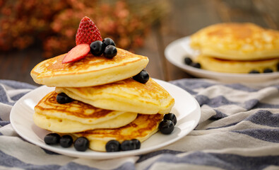 Stack of delicious pancakes sweet food with blueberries and blur dry leaves, plaid fabric in white plate on wooden background.