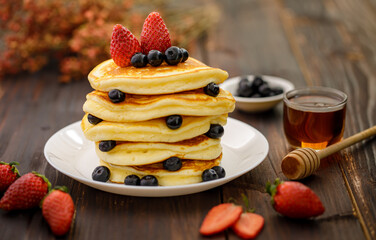 Sweet food. Stack of delicious pancakes with blueberries, Strawberry and honey in white plate on blur wooden background.