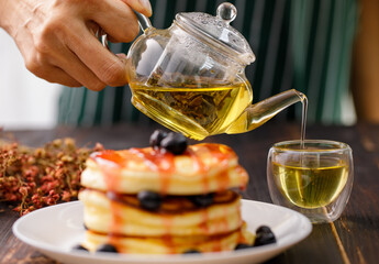 Close up hand pour the tea into the glass. Blur foreground pancakes with strawberry jam, decorated with blueberry fruit in a white plate.
