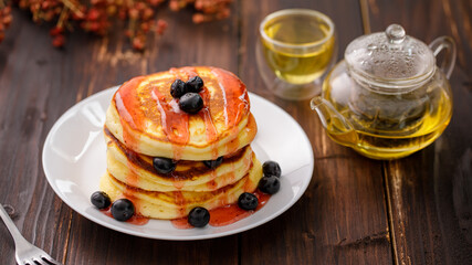 Sweet food. Stack of delicious pancakes with fork rests blueberries and honey, teapot in white plate on blur wooden background.