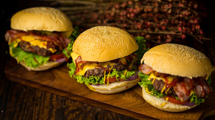 Three hamburgers with beef burger cutlet, fried onion, spinach, ketchup sauce and cheese, served on wood chopping board over dark wooden background.