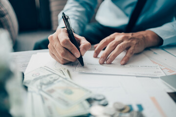 Business man hand writing and signing white blank bank check book and dollar bill, coin, laptop and graph chart on the desk at office. Payment by check, paycheck, payroll concept.