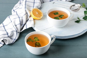 Board with cups of hot tea, parsley and lemon on color wooden background