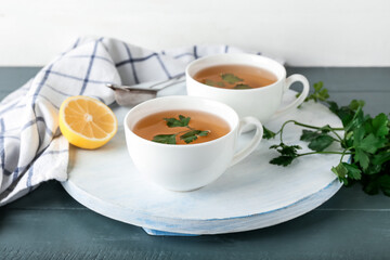 Board with cups of hot tea, parsley and lemon on color wooden background