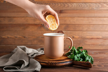 Woman squeezing lemon juice into cup of hot tea with parsley on wooden background