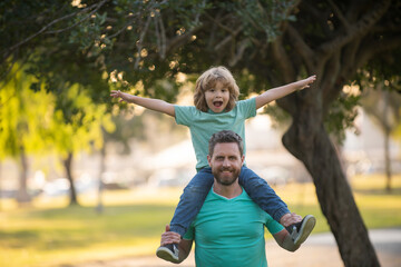 Portrait of a young dad and his child in the park. Happy kid son stretching out hands while his father carrying him on shoulders.