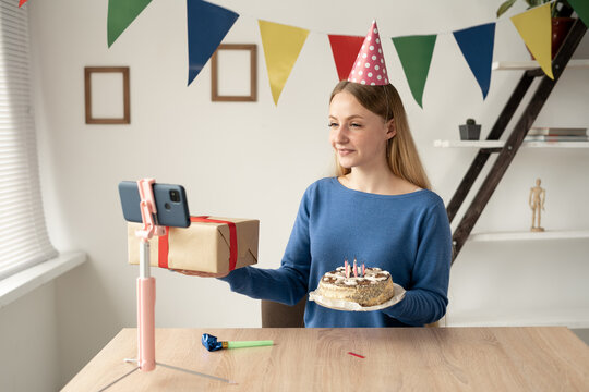 A Smiling Caucasian Blonde Blogger Is Streaming Her Remote Birthday Sitting At A Table Holding A Gift Box And A Birthday Cake. Party In The Office Of A Businesswoman.