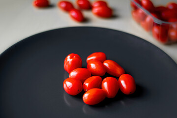 Small tomatoes on a black plate. Red vegetables in a kitchen. Fresh organic food perfect for vegetarian dishes. Selective focus on a tomato, blurred background.