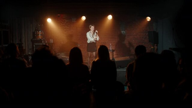 Young Caucasian female comedian entering stage and starting her stand-up monologue inside crowded venue. Shot with ARRI Alexa Mini LF with 2x anamorphic lens