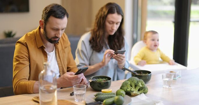 Young parents sitting in smart phones during a lunch time at the table with their one year baby. Dependence of parents on mobile devices and not paying attention to their children