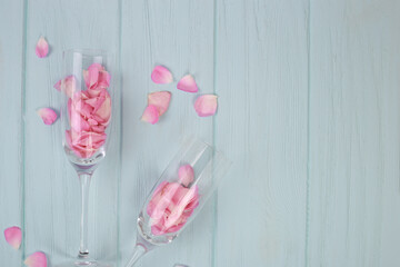 Glasses with flower petals on pastel blue wood background. Romantic atmosphere with pink rose flowers. Date. Valentines day. Wedding. Copy space