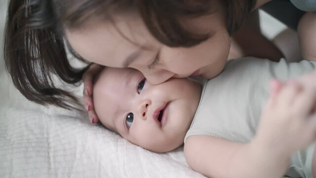 asian woman mother play and kissing baby infant on cheek softly on bed. baby happy when mother parent kissing with love and care. mom using nose touching kiss daughter at home. authentic lifestyle.