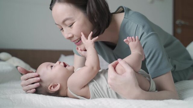 asian woman mother play and kissing baby infant on cheek softly on bed. baby happy when mother parent kissing with love and care. mom using nose touching kiss daughter at home. authentic lifestyle.