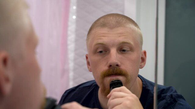 A handsome man with short hair shaves his beard with a trimer while standing in the bathroom in front of the mirror. Daily routine of personal care.