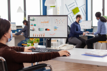 Disabled handicapped businesswoman with face mask immobilized in wheelchair working at financial project typing on pc showing data, prosessing on display sitting in new normal startup business office.