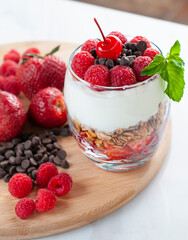 A glass of yogurt with raspberries granola, strawberries and chocolate chips with a mint or peppermint leaf on top over a wooden cutting board in a white background