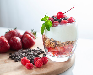 A glass of yogurt with raspberries granola, strawberries and chocolate chips with a mint or peppermint leaf on top over a wooden cutting board in a white background lit by a window