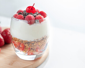 A glass of yogurt with raspberries granola, strawberries and chocolate chips  over a wooden cutting board in a white background with strawberries  on the side, horizontal close up with window light.
