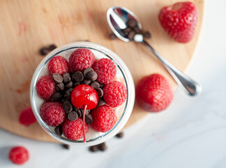 A glass of yogurt with raspberries granola, strawberries and chocolate chips with a spoon  over a wooden cutting board in a white background