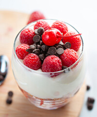 A glass of yogurt with raspberries granola, strawberries and chocolate chips with a spoon on the side over a wooden cutting board in a white background