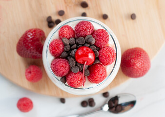 A glass of yogurt with raspberries granola, strawberries and chocolate chips with a spoon on the side over a wooden cutting board in a white background, horizontal.
