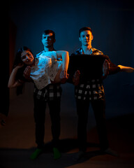 Two guys in a plaid shirt are holding a girl on a dark background in a studio with multicolored light.