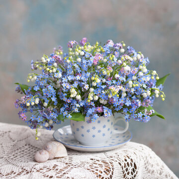 A bouquet of spring flowers forget-me-nots and lilies of the valley in a mug on a table with a lace tablecloth against the background of a colorful wall. Beautiful card.