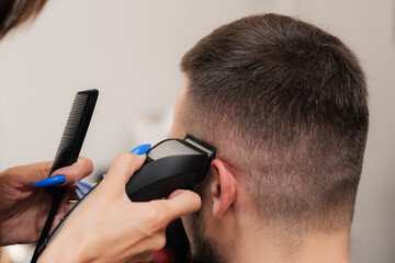 A female barber cuts the hair of a young man with a clipper. Close-up