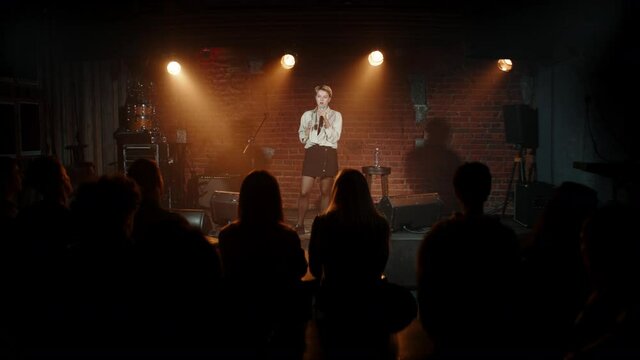 Young Caucasian female comedian performing her stand-up monologue on a stage of a small venue. Shot with ARRI Alexa Mini LF with 2x anamorphic lens