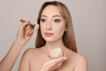 Professional makeup artist applying powder onto beautiful young woman's face with brush on grey...
