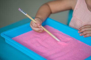 Little girl drawing letter on colored pink sand indoor.