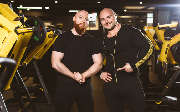 Portrait Of Couple Of Two Muscular Men. Team Of Trainers In The Same Clothes Posing For The Camera Against The Background Of The Gym