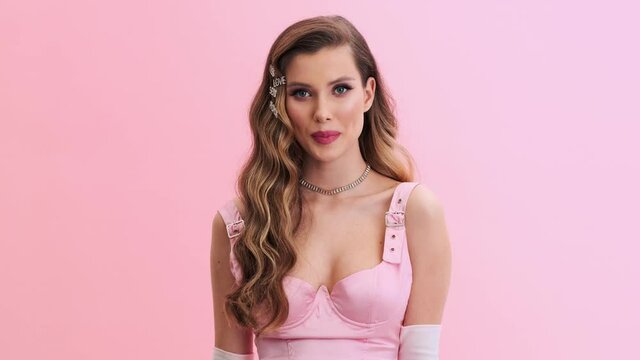 A beautiful young woman wearing gorgeous evening pink dress with gloves and hairpins is posing to the camera standing isolated over a pink wall in the studio