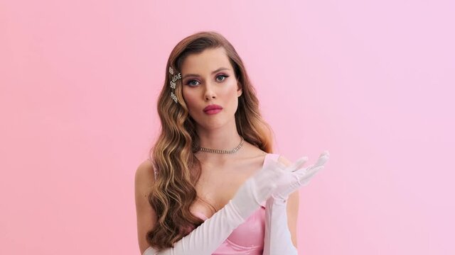 A displeased young woman wearing gorgeous evening pink dress with gloves and hairpins is clapping her hands standing isolated over a pink wall in the studio