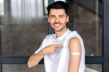 Obraz premium Happy healthy young Caucasian male, in casual clothes, received a vaccine against covid 19, immunity from viruses and infections, take care of his health, points to a band-aid on his shoulder, smiling