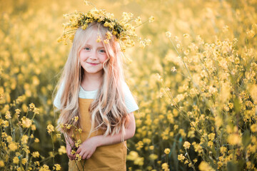 Smiling blonde beautiful child girl 4-5 year old wear floral wreath and summer stylish clothes stand in flower field outdoors. Happy toddler over nature background close up. Looking at camera.