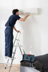 young handyman in overalls standing on ladder while fixing broken air conditioner