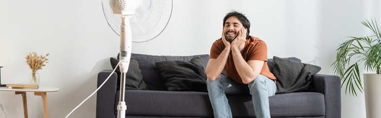 happy bearded man sitting on couch near blurred electric fan, banner