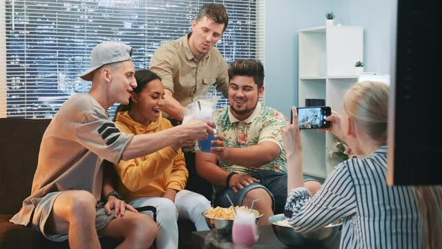 Close up of girl making photo of friends sitting on sofa with cocktails by smartphone