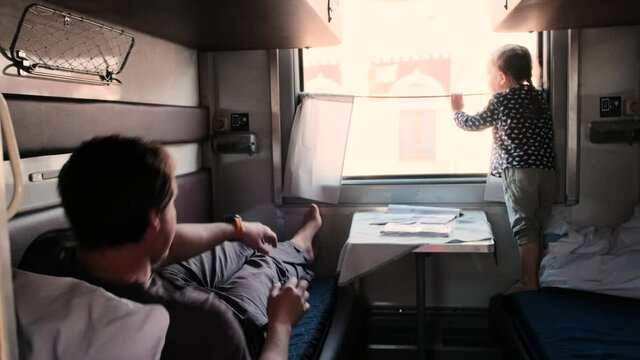 Little daughter in dot shirt looks out of window while father lies on bed and picks up phone from table in train sleeping car