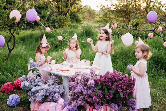 Summer outdoor kids birthday party. Group of happy Children celebrating birthday in park. Children blow candles on birthday cake. Kids party lilac pink pastel decoration and food. Presents and sweets.