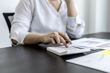 A businesswoman is pressing a white calculator, she is calculating the numbers on the company financial information sheet to verify the accuracy before attending a meeting with a business partner.