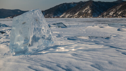Transparent shiny shard of ice close-up. Sun glare on the edges, shadow on the snow. Background -...